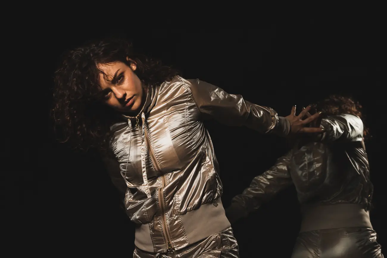 Frau in silbernem glänzenden Anzug posiert seitlich gegen schwarzen Hintergrund; linke Arm ausgestreckt, rechte Arm hinten; leicht reflektierende Oberfläche.